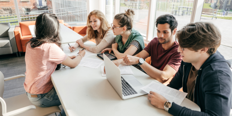 Four students engaged in a discussion at a table, each using a laptop, focused on their pharmacy and healthcare studies.