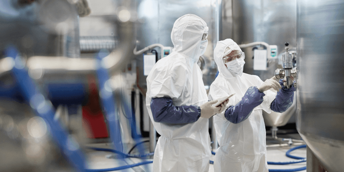 Two individuals in protective gear conducting quality control tests in a pharmaceutical factory setting.